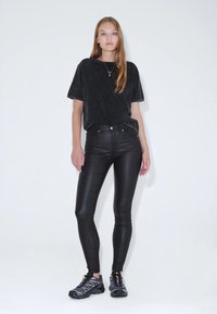 Černé tričko s krátkým rukávem a měkkou texturou, spárované s těsnými černými kalhotami z faux kůže. Obuv tvoří černé sportovní boty s barevnými akcenty.