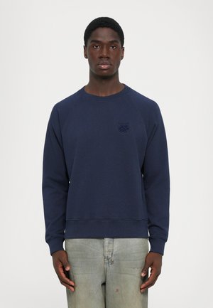 Man wearing a navy blue sweatshirt with a small chest logo and faded light gray jeans, standing against a plain white background.