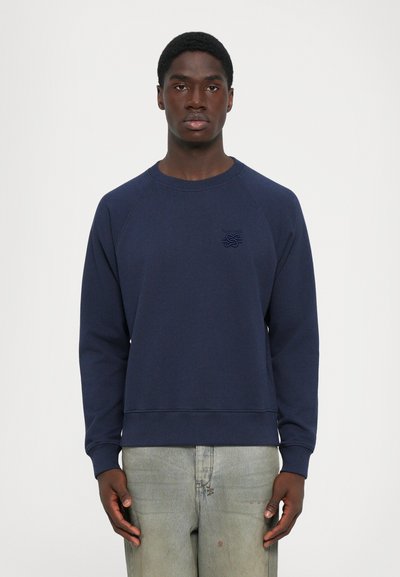 Mand iført en marineblå sweatshirt med et lille brystlogo og falmede lysegrå jeans, stående foran en ensfarvet hvid baggrund.