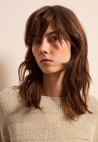 Beige gestrickter Pullover mit loser Maschenstruktur und weitem Ausschnitt, getragen von einer Person mit zerzaustem braunem Haar und neutralem Hintergrund.