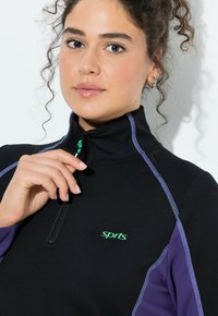 Mladá žena s kudrnatými vlasy si upravuje zip na černo-fialovém sportovním topu se zeleným logem "sprts", bílý pozadí.