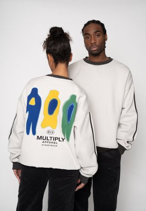 To modeller iført hvide sweatshirts med sorte manchetter og sorte bukser; den ene vender fremad, den anden viser et farverigt abstrakt design på bagsiden.