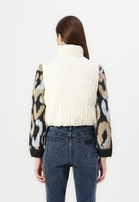 Προστατευτικό γιλέκο με ψηλό γιακά σε κρεμ πλέξη, που διαθέτει μανίκια με λεοπάρ σχέδιο σε καφέ και μαύρο χρώμα. Φοριέται πάνω από σκούρες τζιν παντελόνες.