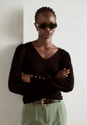 Black V-neck sweater with long sleeves and button accents at cuffs, paired with green trousers and a black belt featuring a gold buckle.