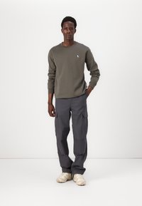 Olijfgroene crewneck sweatshirt met subtiele vogellogo; gecombineerd met grijze cargobroek met zijzakken; beige sneakers.