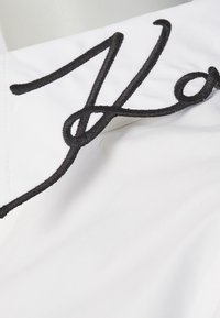 Vitt tyg med svart broderad skrivdetalj, som har en slät textur och en modern kursiv design.