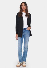 Črn kardigan na odprto preko belega majice, v kombinaciji s svetlo modrimi kavbojkami in kremnimi supergami. Ima sproščen kroj in preprost dizajn.