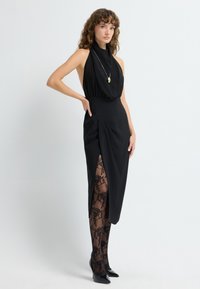 Czarna sukienka bez rękawów z wysokim dekoltem, rozcięciem po bokach i drapowaniem. W komplecie przezroczyste czarne rajstopy z kwiatowym wzorem oraz czarne szpilki.