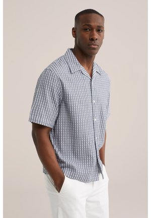 Moški v kratkemu rokavu z vzorčasto srajco z ovratnikom in belimi hlačami, stoji z rokami v žepih pred enobarvnim ozadjem.