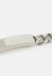 Ασημένια βραχιόλι αλυσίδας με λείο, ανάγλυφο μπάρα που φέρει τη λέξη "GUESS" και σχέδιο αλυσίδας με διασύνδεση. Λαμπερή επιφάνεια με μεταλλική γυαλάδα.