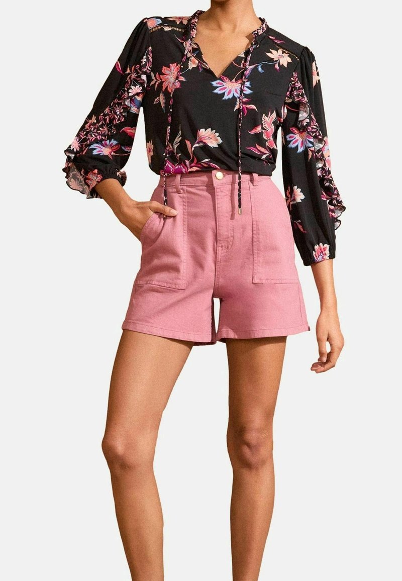 Blusa nera a fiori con maniche lunghe, abbinata a pantaloni corti rosa chiaro a vita alta con tasche frontali e una texture liscia.