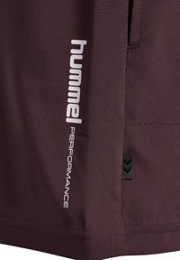 Mørk rødbrun stof med hvid lodret "hummel PERFORMANCE" tekst og et lille sort mærke med to vinkler.