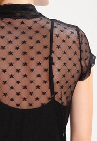 Top de malla negro con un diseño transparente y patrones de estrellas, mangas cortas y cuello redondo, que ofrece una apariencia texturizada y ligera.