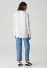 Weißes Langarmshirt mit abgerundetem Saum, kombiniert mit hellblauen geraden Jeans und marineblauen Sneakers mit weißen Akzenten.