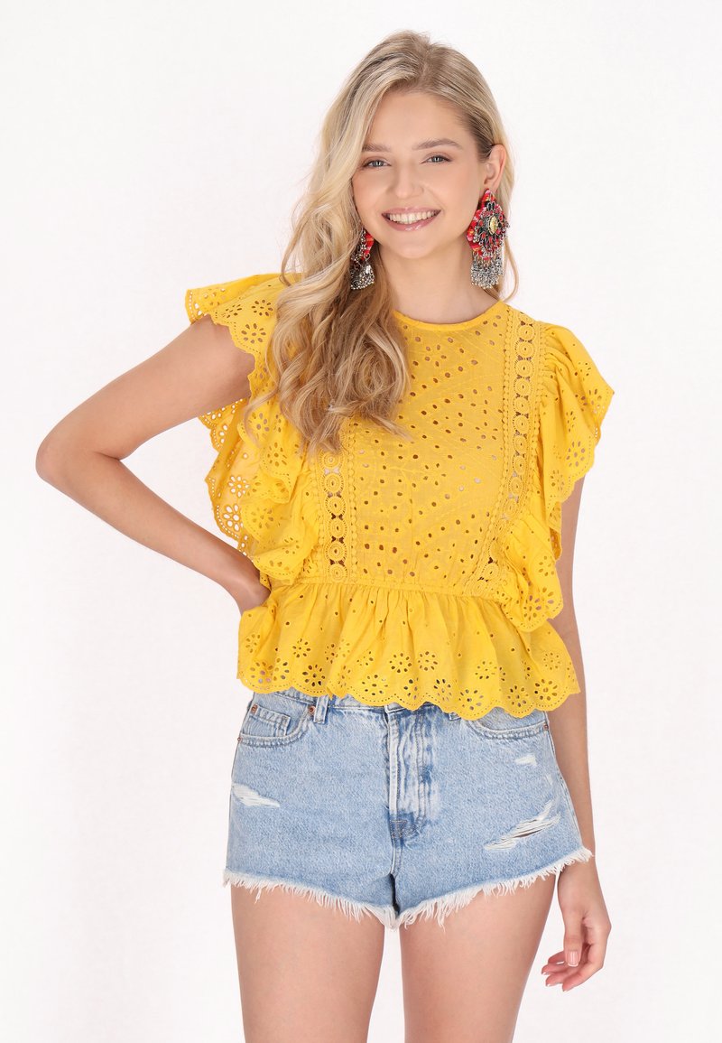 Top giallo in pizzo con orlo a frange, maniche corte e bordi scallop, abbinato a pantaloncini di jeans strappati.