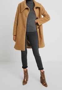 Persona che indossa un cappotto teddy color cammello, un maglione grigio a collo alto, jeans scuri arrotolati e stivaletti alla caviglia in pelle di serpente, in piedi su un pavimento bianco.