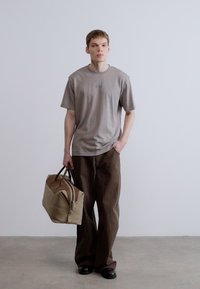 Szürke pamut póló, laza szabás, barna bő szárú nadrággal párosítva. Bézs duffel táska barna díszítésekkel, a bal kézben tartva. Lapos talpú cipő.