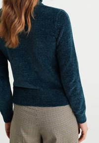 Mörk turkos stickad tröja med hög hals och ribbade manschetter, kombinerad med bruna rutiga byxor. Texturen verkar mjuk och lätt skimrande.