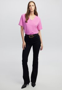 Roze v-hals t-shirt gecombineerd met zwarte uitlopende jeans en een zwarte riem met een gouden gesp. Het model draagt zwarte puntige hakken. Soepele stof.