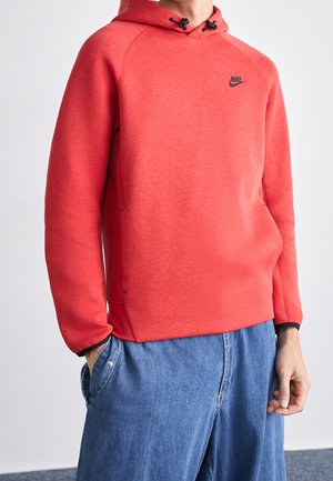 Person trägt roten Nike-Hoodie mit schwarzem Logo, Hände in den Taschen, kombiniert mit locker sitzenden blauen Denim-Jeans vor einfarbigem Hintergrund.