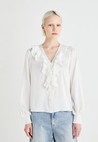 Blusa bianca con davanti arricciato, scollo a V e maniche lunghe con polsini elasticizzati. Abbinata a jeans in denim chiaro con effetto usurato.