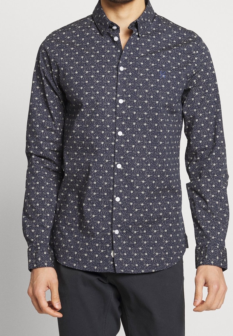 Homme portant une chemise bleu marine boutonnée avec un petit motif floral blanc et un pantalon foncé, debout devant un fond uni.