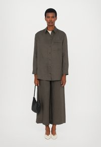Persona che indossa una camicia verde oliva con bottoni e pantaloni larghi, tiene in una mano una borsa nera intrecciata, scarpe bianche a punta su uno sfondo bianco.