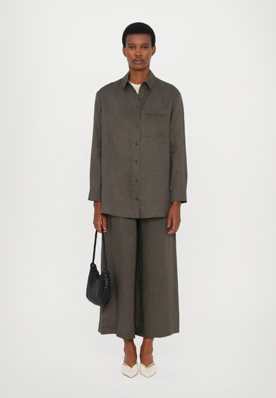 Άτομο που φοράει ένα ελαιόχρωμο πουκάμισο με κουμπιά και φαρδιά παντελόνια, κρατώντας μία μαύρη πλεκτή τσάντα στο ένα χέρι, με λευκές μυτερές παπούτσια σε άσπρο φόντο.