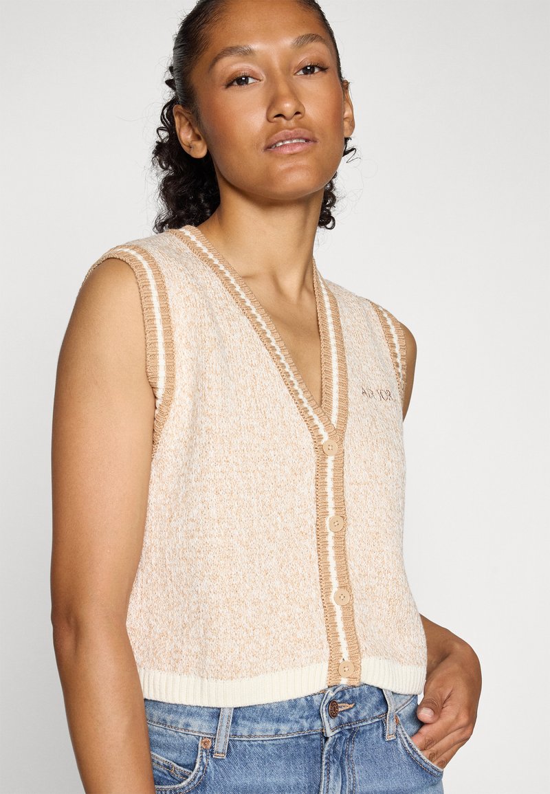 Ärmellose Strickjacke aus strukturiertem beigefarbenem Material mit V-Ausschnitt, kontrastierendem weißen Besatz und fünf cremefarbenen Knöpfen auf der Vorderseite.