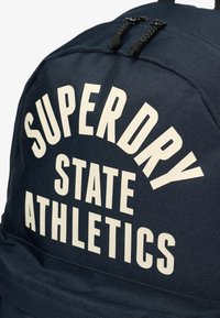 Sac à dos bleu marine avec un grand texte blanc indiquant "SUPERDRY STATE ATHLETICS", avec des fermetures éclair noires et un tissu texturé.