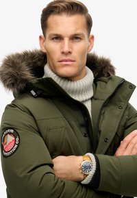 Parka verde oliva con capucha de piel sintética, parche de marca prominente en la manga, combinada con un suéter de canalé gris claro y un reloj dorado.