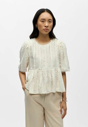 Femme portant un chemisier crème à manches courtes texturé avec des rayures verticales froncées et un pantalon beige à jambes larges, debout devant un fond blanc.