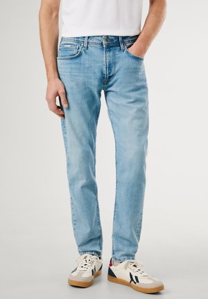 Personne portant un jean slim bleu clair, des baskets blanches avec des détails noirs et rouges, et un t-shirt blanc, debout avec une main dans la poche.