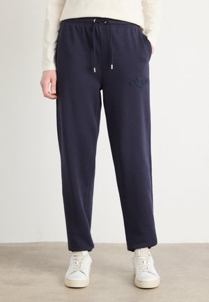 Persoană purtând pantaloni de trening bleumarin cu șnur și buzunare laterale și adidași albi, stând pe un podea bej.