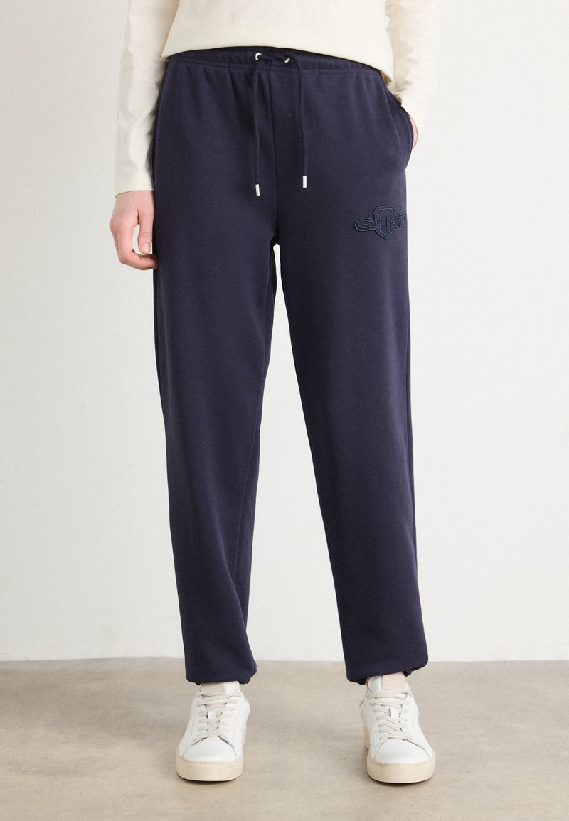 Pantaloni da tuta blu navy con una vita regolabile con cordino, gambe affusolate e dettagli del logo ricamato. Abbinati a sneakers bianche. Texture liscia.