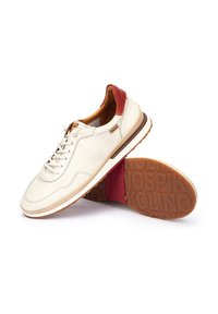 Zapatillas de cuero blancas con acentos rojos, con una suela texturizada en tonos marrones, cierre de cordones y un interior acolchado. Logo de la marca visible en el lateral.