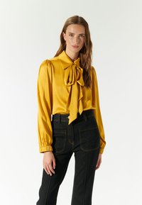 Blusa di raso gialla con un ampio colletto a fiocco, maniche a sbuffo e polsini elasticizzati, abbinata a jeans in denim scuro a vita alta con ricami.