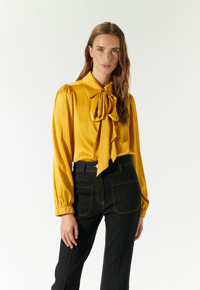 Blusa di raso gialla con un ampio colletto a fiocco, maniche a sbuffo e polsini elasticizzati, abbinata a jeans in denim scuro a vita alta con ricami.
