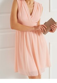 Světle růžové šaty s draped výstřihem, plisovanou částí bodice a splývavou sukní. V jedné ruce drží kabelku s zlatými akcenty.