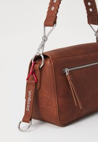 Bolso de cuero marrón con patrones florales en relieve, herrajes plateados y una correa trenzada con un acento rojo y etiqueta de marca.