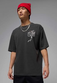 Black cotton t-shirt with short sleeves, featuring a graphic basketball motif on the chest. Worn with a red beanie and silver chains.