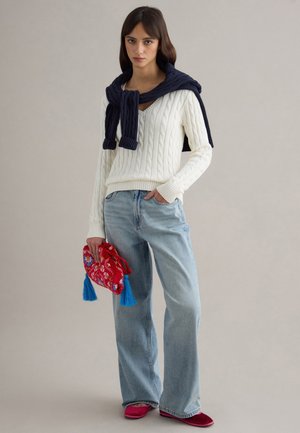 Mujer con suéter blanco de punto grueso, jeans azul claro, suéter azul marino sobre los hombros, sosteniendo un bolso rojo bordado con borlas azules.