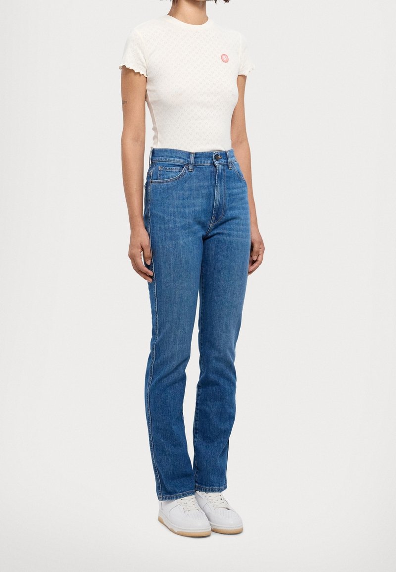 Bílý texturovaný top s krátkým rukávem a vlnitým lemem, doplněný malým růžovým logem, v kombinaci s rovnými modrými džíny a bílými teniskami.