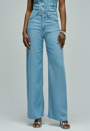 Persona con vaqueros acampanados de tiro alto azul claro y tacones plateados con tiras, de pie con los brazos relajados a los lados sobre fondo gris.