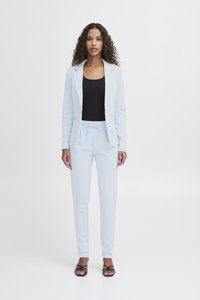 Žena s kovrčavom kosom nosi svijetloplavi sako i hlače, crnu majicu i crne sandale s remenčićima, stojeći nasuprot bijele pozadine.