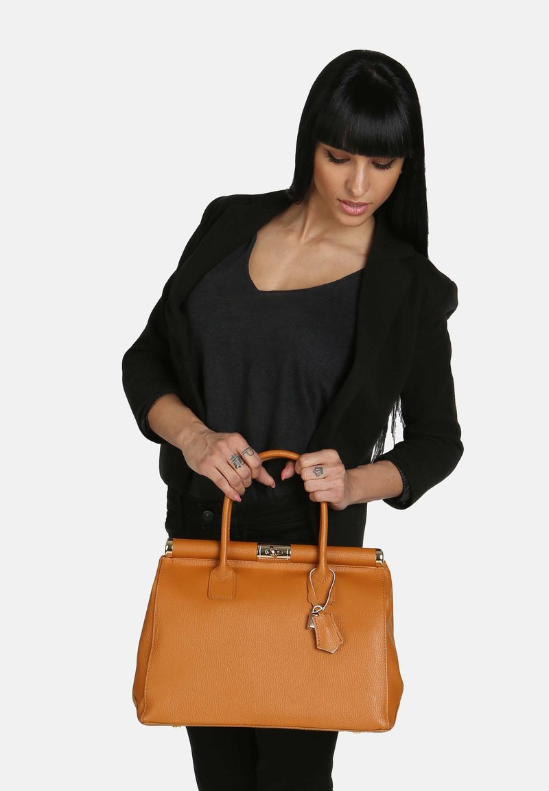 Tan läderhandväska med en strukturerad form, topphandtag och metalliska accenter. Har en texturerad yta och en etikett hängande från sidan.