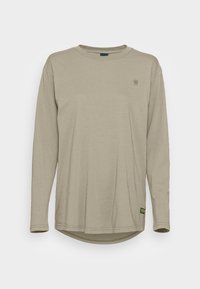 Beige långärmad t-shirt med rund halsringning, med en liten logotyp på vänster bröst och en svart och gul etikett nära nederkanten.