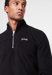Maglione in pile nero con zip a quarto. Presenta un logo "Schott" rosso e bianco sul petto. Texture morbida e collo alto.