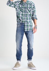 Camisa de cuadros verde y blanca de manga larga con dos bolsillos en el pecho, combinada con jeans azul claro y zapatillas beis, con los puños arremangados.