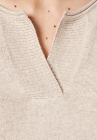 Beige Strickpullover mit tiefem V-Ausschnitt und gerippten Akzenten, weicher Textur und subtilen Fäden im gesamten Stoff.